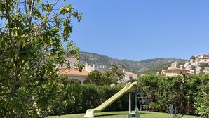 Un parque infantil con tobogán y columpios, rodeado de césped verde y árboles, con casas y colinas al fondo.