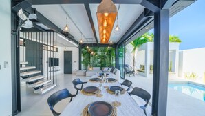 A modern dining area with a long table set for six, surrounded by black chairs, with a pool visible through large windows. - Villa Bali Townhouse (Kerobokan)