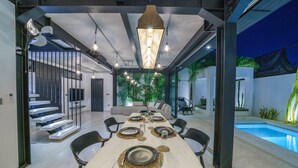 A modern dining area with a long table set for six, surrounded by chairs, and a pool visible through the glass walls. - Villa Bali Townhouse (Kerobokan)