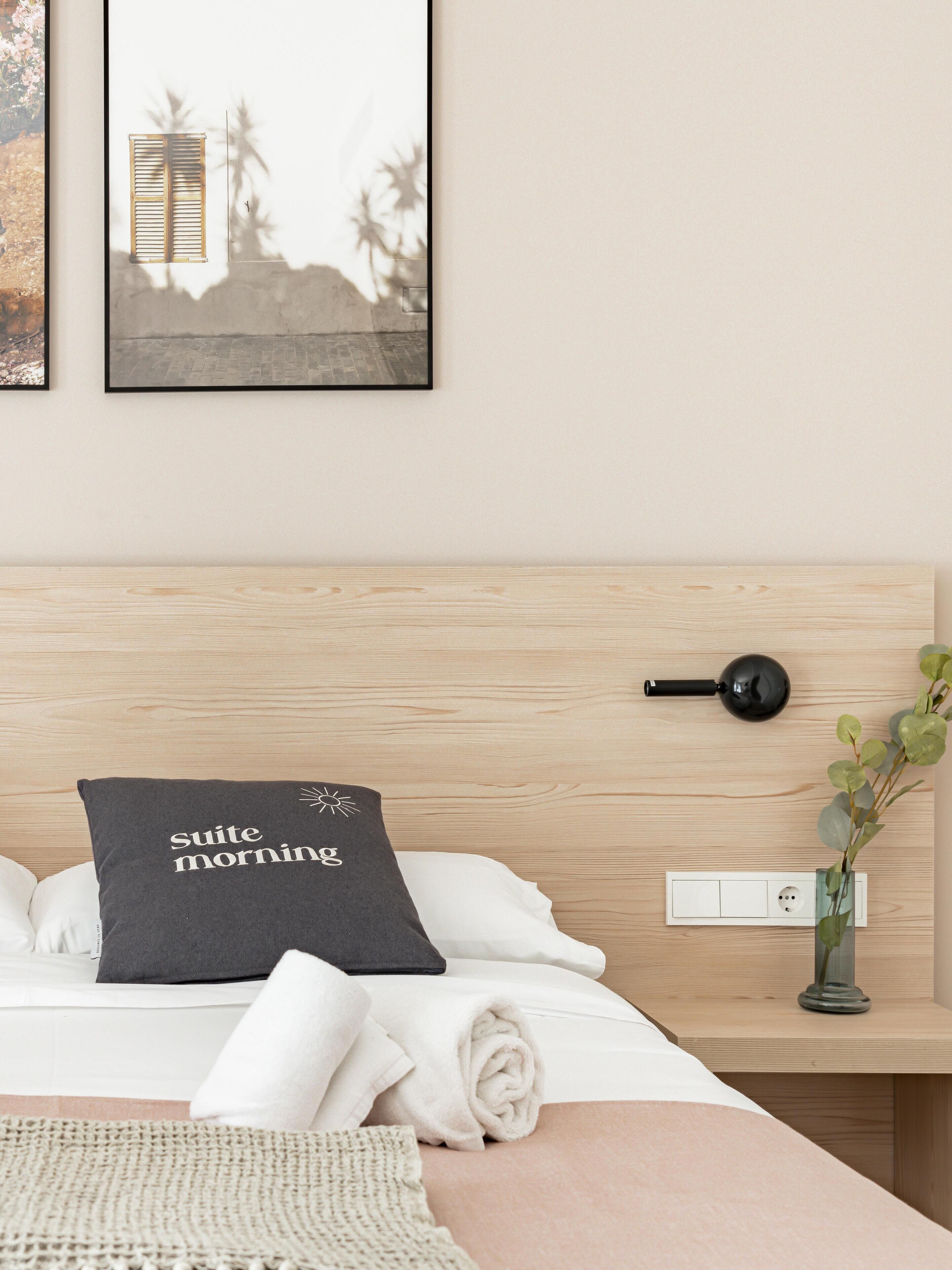 A neatly made bed with a pillow that reads "Suite morning," a bedside table with a black lamp, and a wall-mounted power outlet.