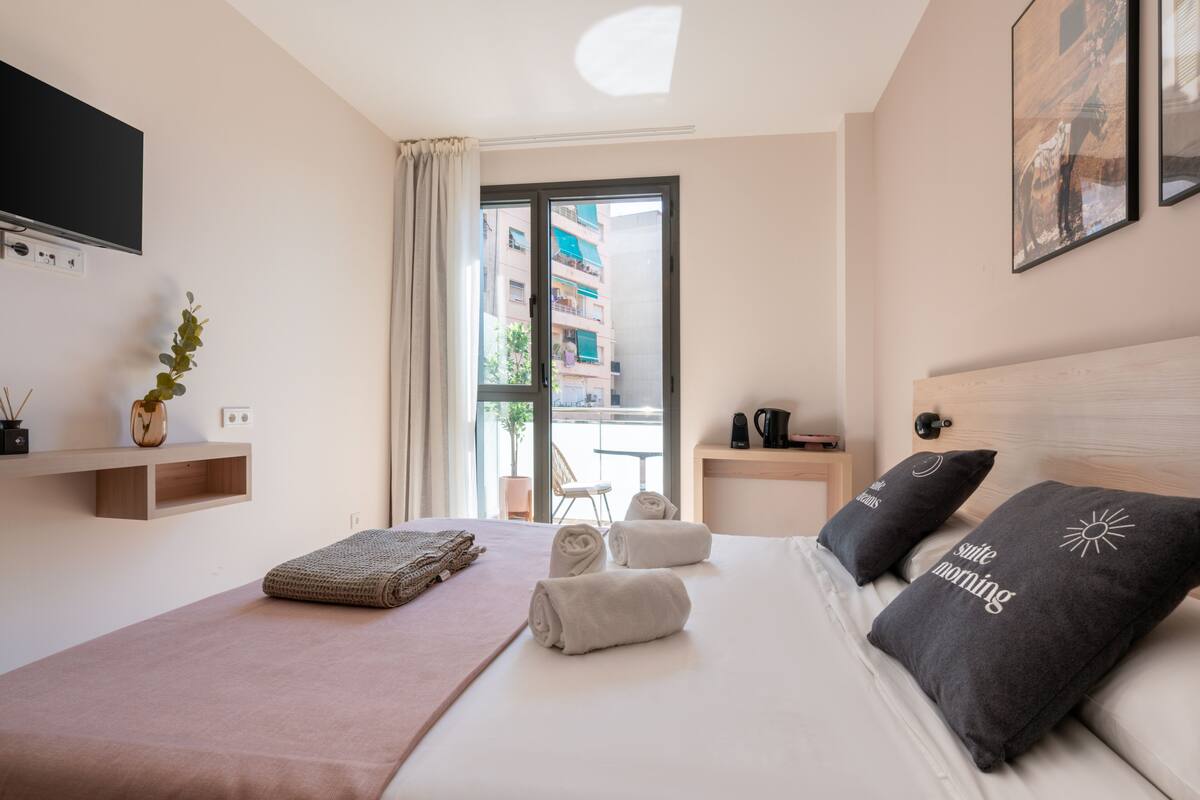 A modern hotel room with a large bed, a flat-screen TV, a small table with a coffee maker, and a window with a view of the city.