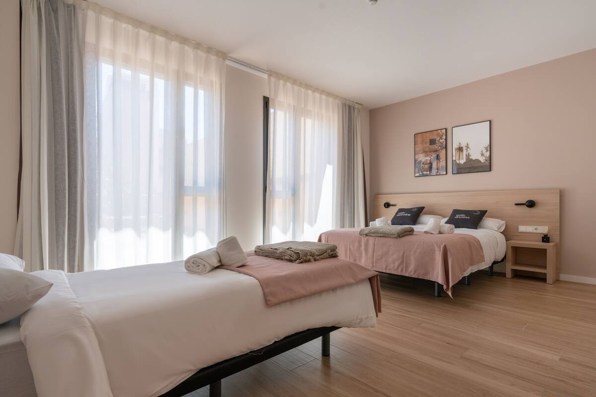 A hotel room with two beds, a wooden floor, and a large window with sheer curtains.