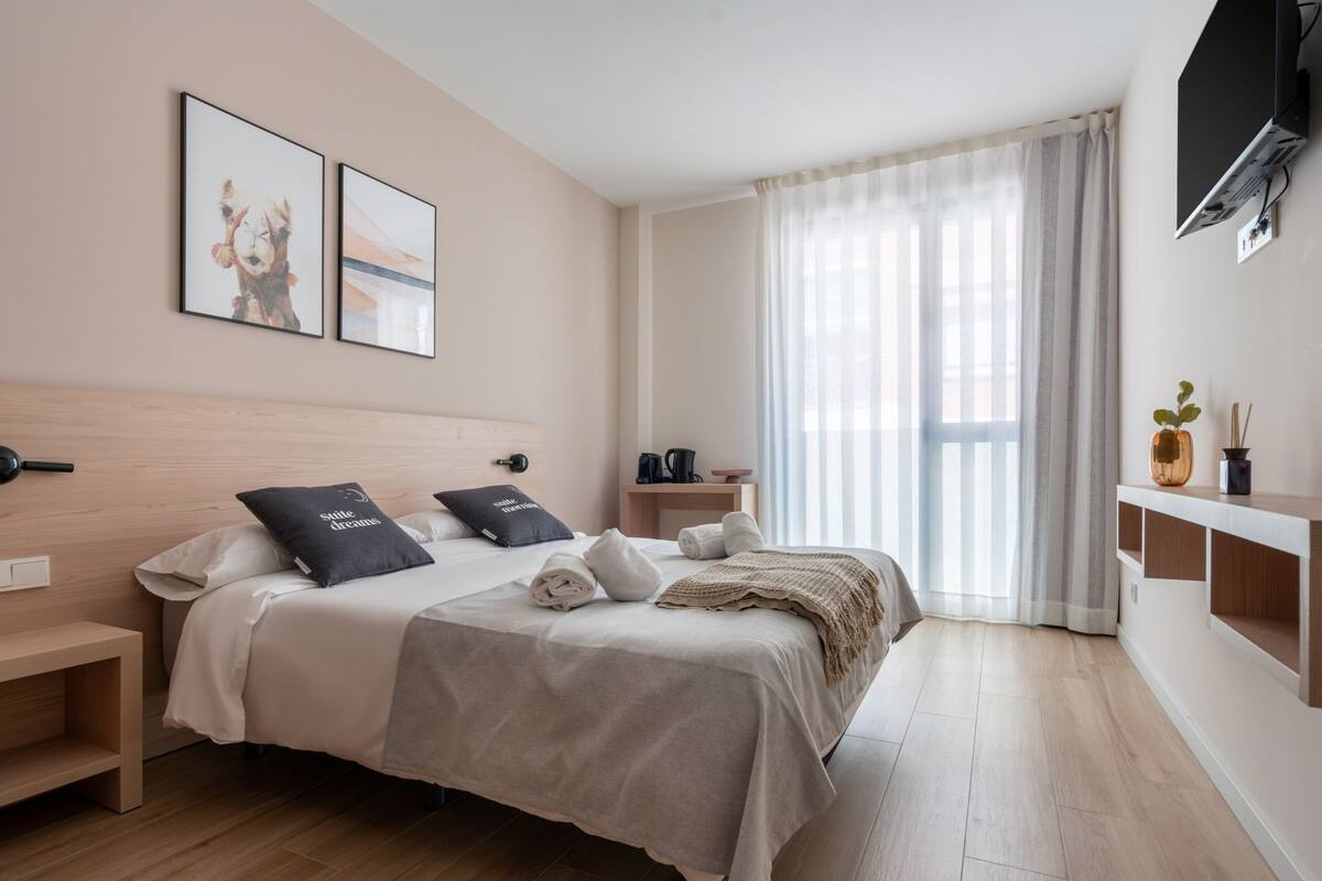 A modern hotel room with a large bed, a bedside table, a TV mounted on the wall, and a window with sheer curtains.