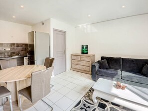 A modern living room with a grey sofa, a wooden dining table with chairs, a kitchen area with a fridge, and a white door.