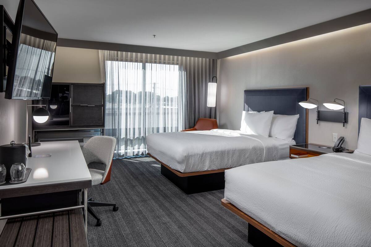 A hotel room with two beds, a desk, a chair, a TV, and a large window with curtains.