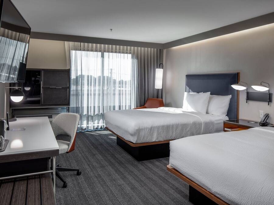 A hotel room with two beds, a desk, a chair, a TV, and a large window with curtains.