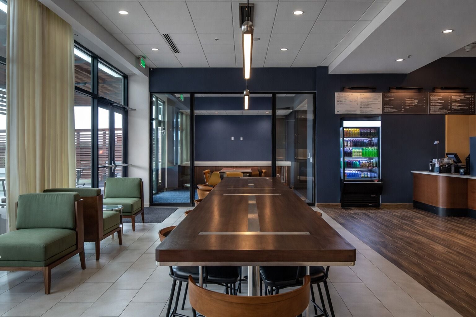 A modern hotel lobby with a long wooden reception desk, comfortable seating, a refrigerator with beverages, and a view through the glass doors.