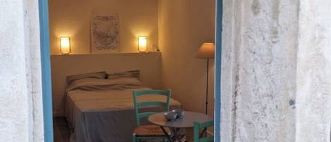 A small, rustic room with a bed, two chairs, a table, and two wall-mounted lamps.