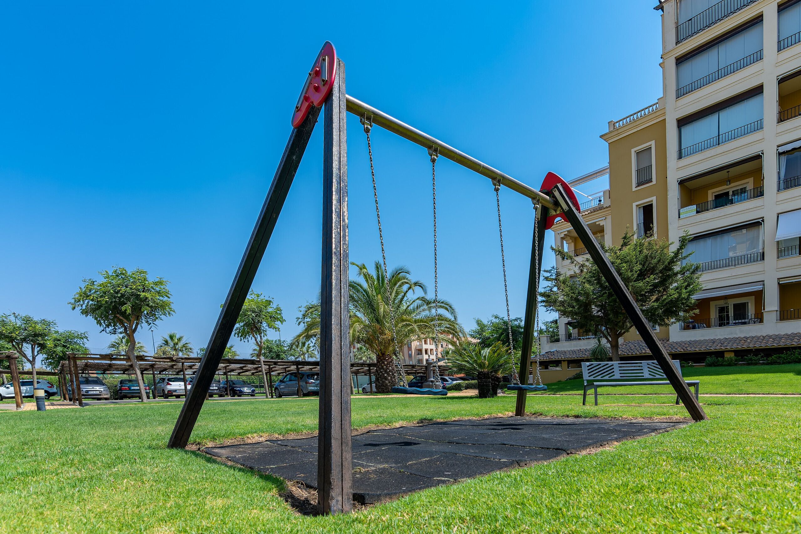 Un parque infantil con columpios y un banco, rodeado de césped y palmeras, con un edificio de varios pisos al fondo.