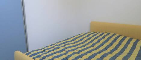 Una cama con cabecera amarilla y colcha azul y amarilla en una habitación con pared blanca lisa y suelo gris.