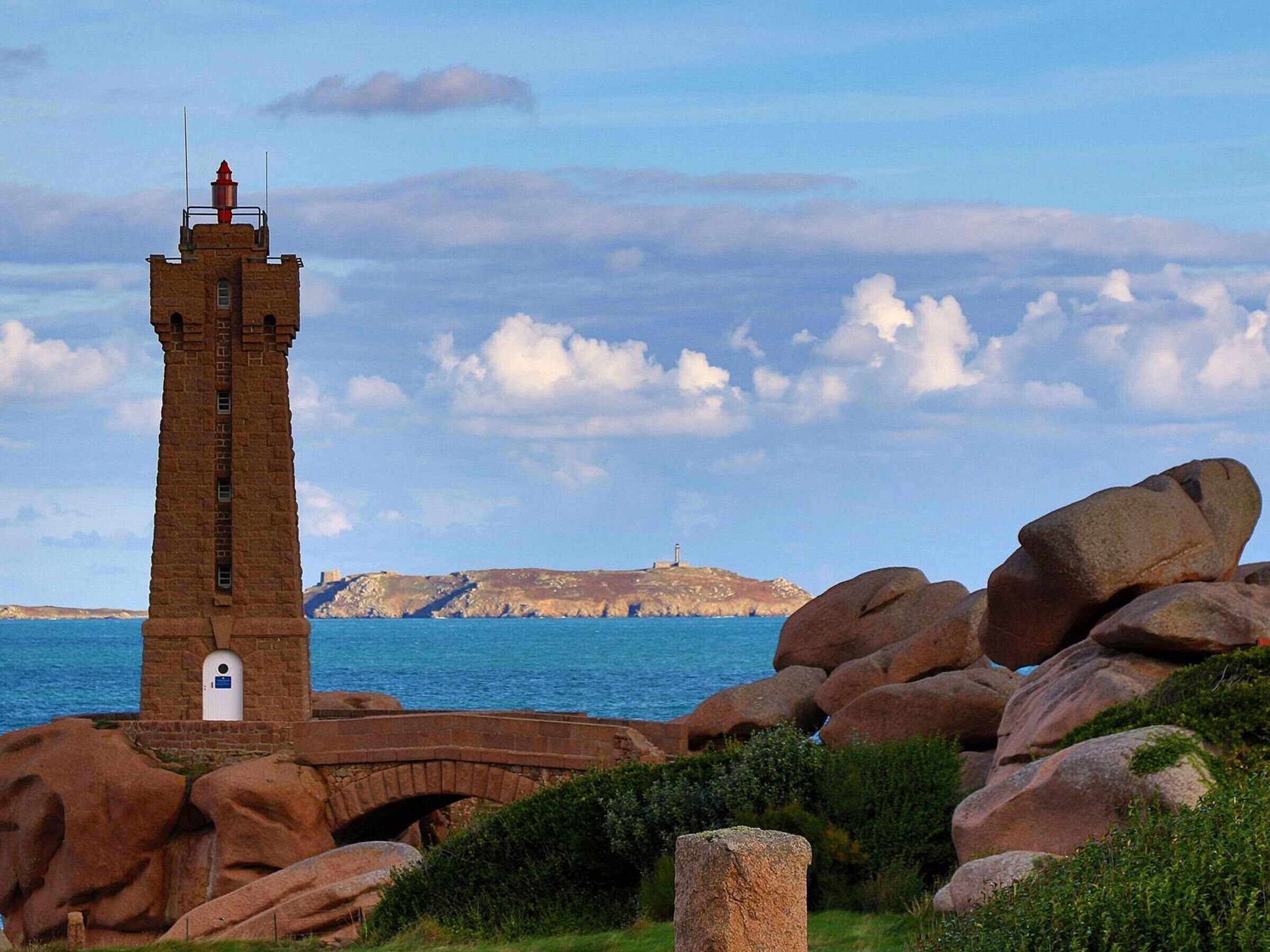 Un faro con torre de ladrillo, un puente de piedra y grandes rocas en un entorno costero.