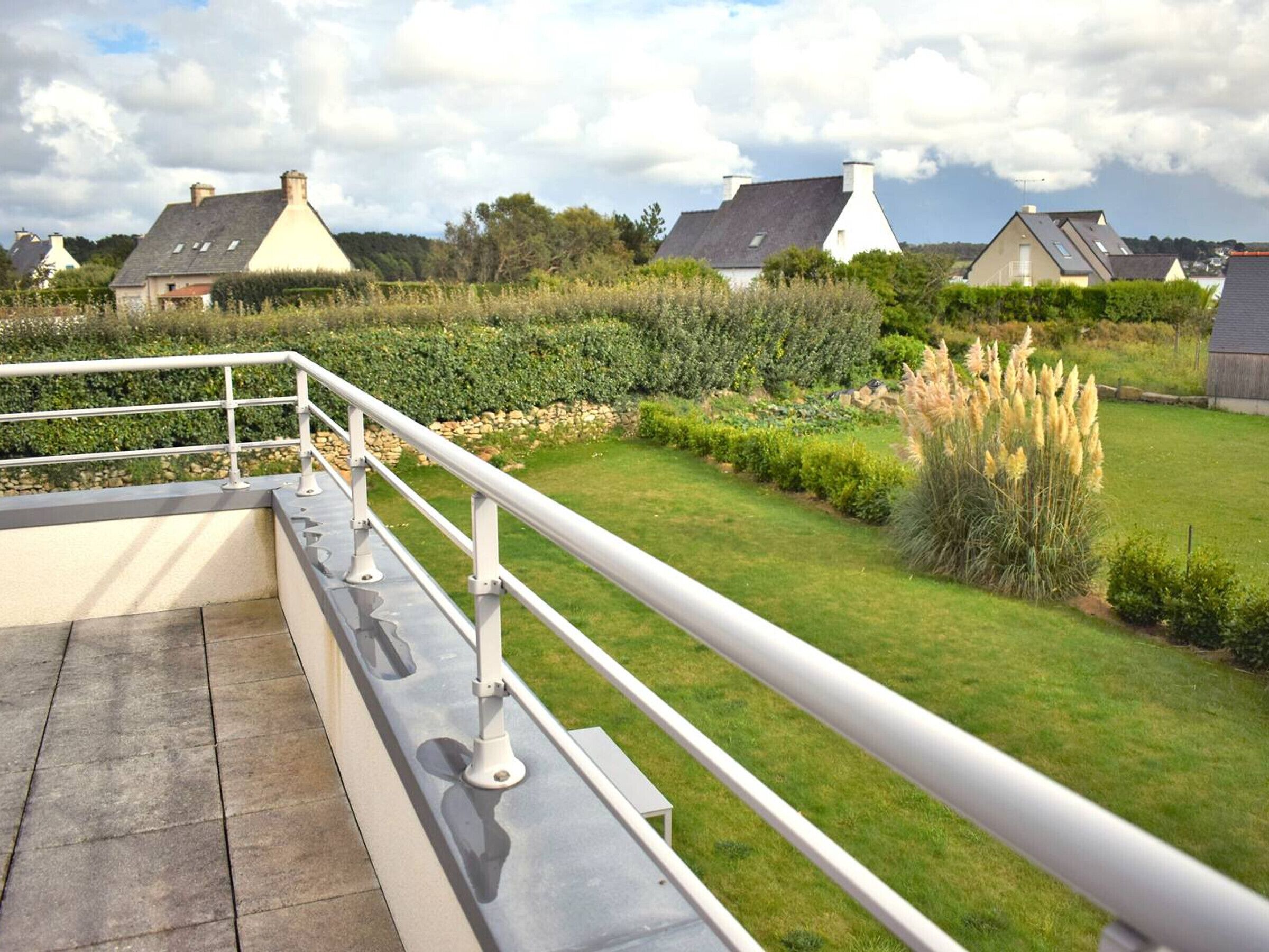 Una terraza con barandilla de metal, suelo de piedra y vistas a casas y vegetación.