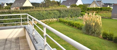 Una terraza con barandilla de metal, suelo de piedra y vistas a casas y vegetación.