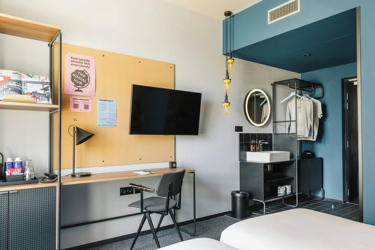 Een hotelkamer met een bureau, stoel, bed, televisie en een badkamer met wastafel en spiegel.