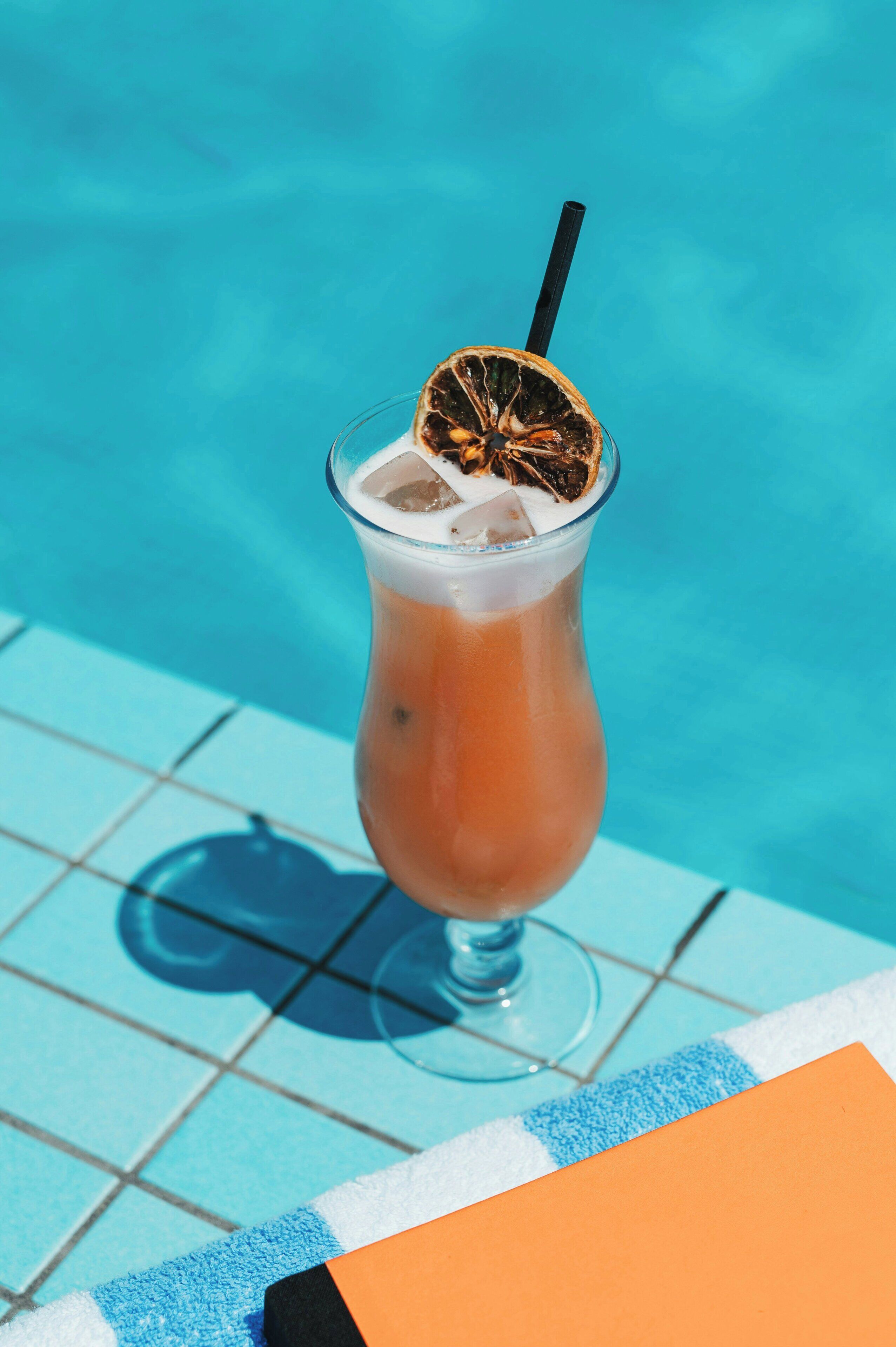 A cocktail with a straw and a slice of fruit in a glass, placed on a poolside area with tiled flooring.