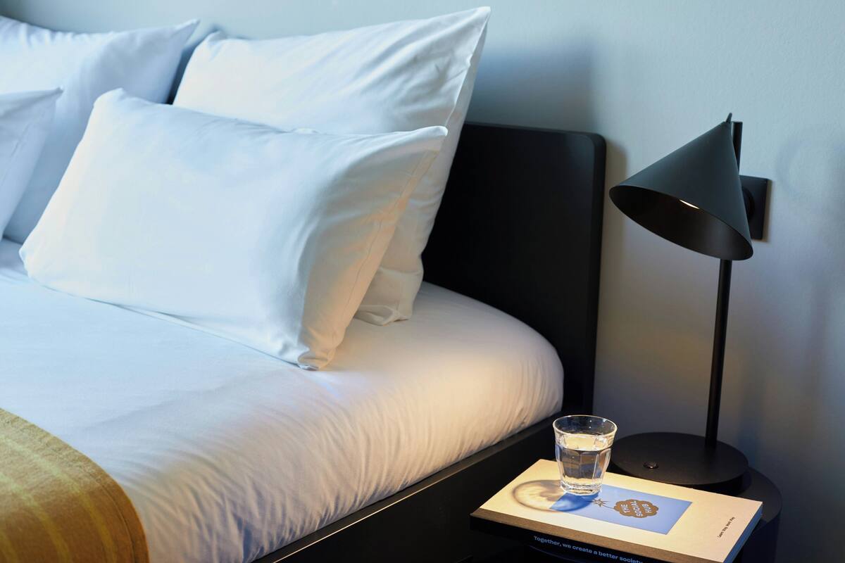 Een netjes opgemaakt bed met witte lakens, een zwarte vloerlamp en een glas water op een nachtkastje met een boek.