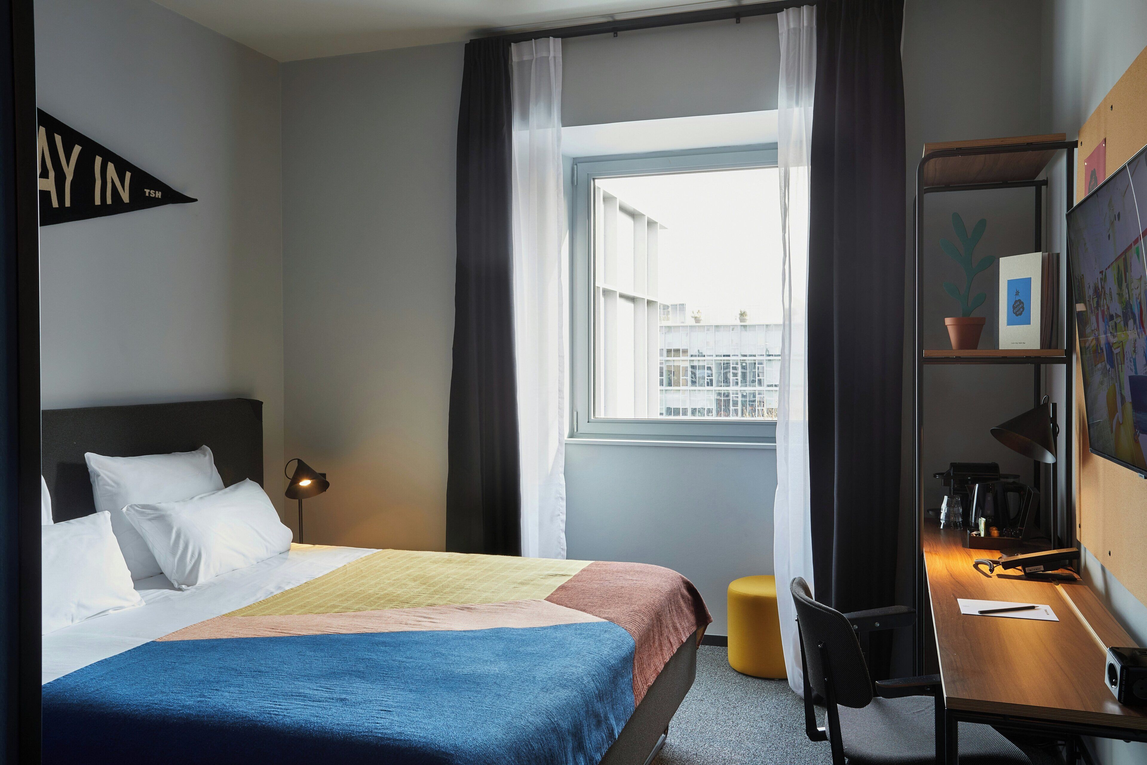 A hotel room with a bed, a desk, a chair, a lamp, a window with curtains, and a wall-mounted shelf with a plant.