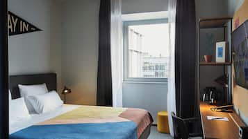 Habitación de hotel con cama, escritorio, silla, lámpara, ventana con cortinas y estante de pared con una planta.