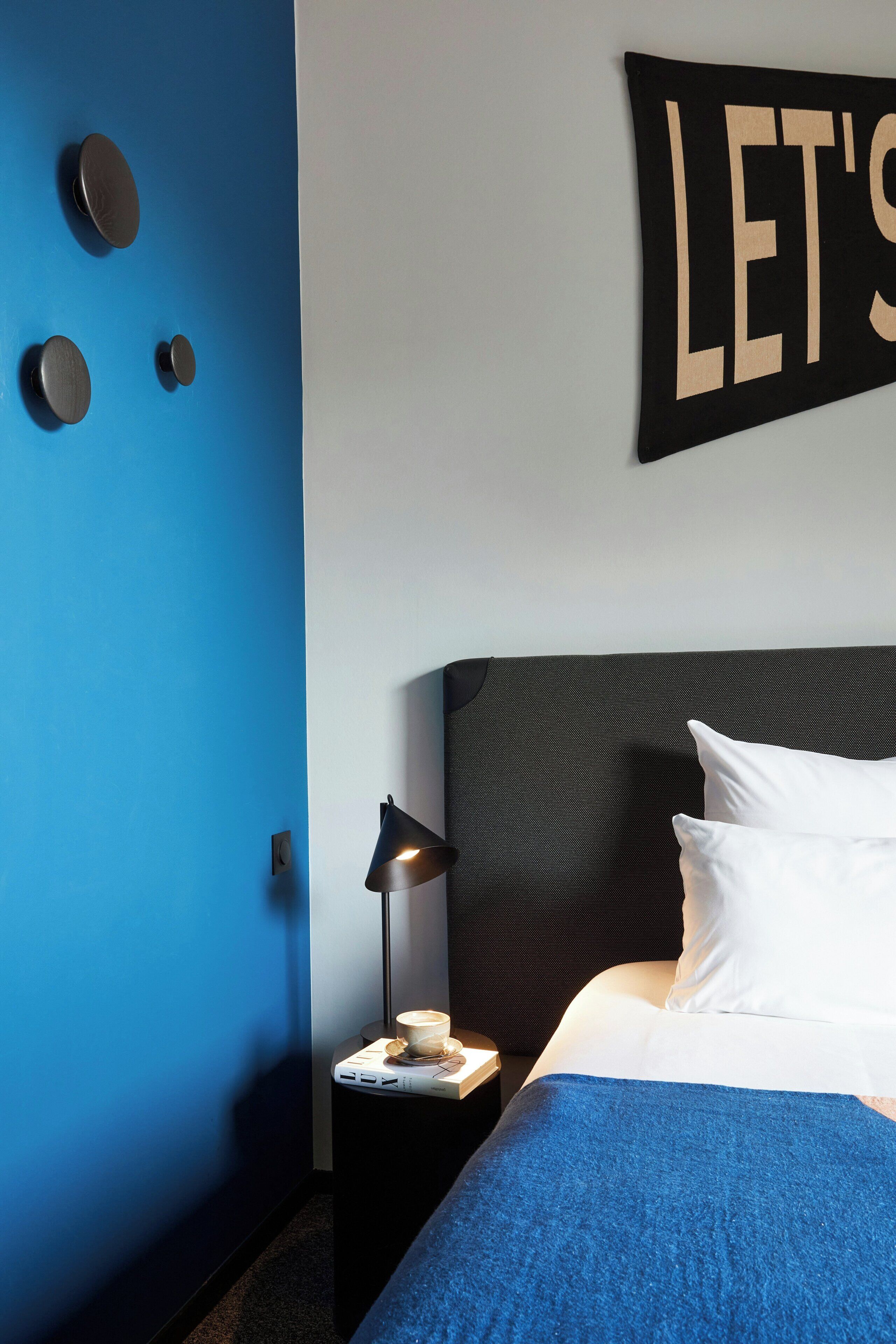 A hotel room with a blue wall, a bed with white pillows, a black headboard, a nightstand with a lamp, and a sign that says 'LET'S'.