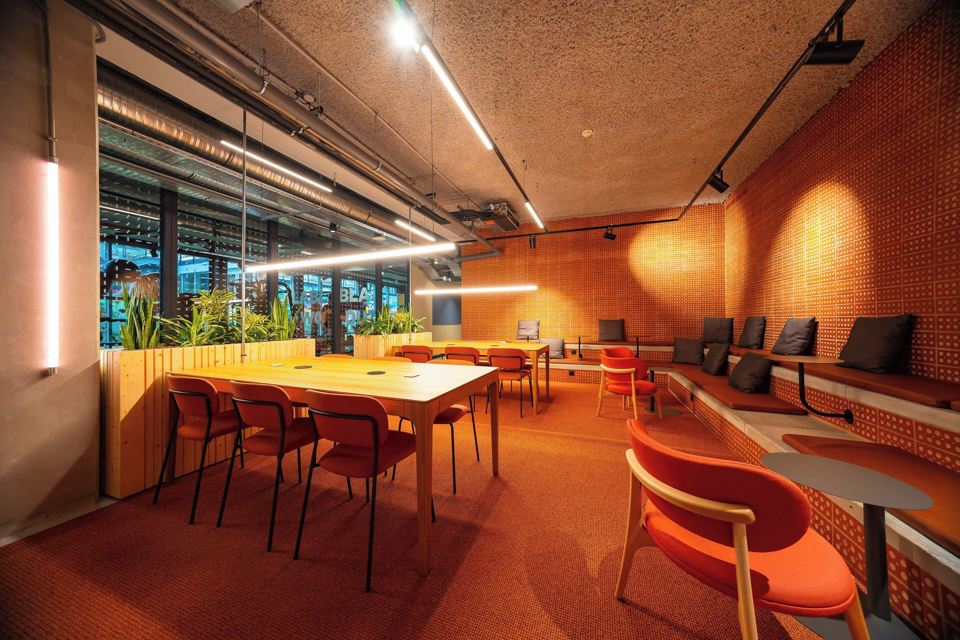 Un espacio interior con una mesa larga, varias sillas, una zona de estar con bancos y un muro con diseño.