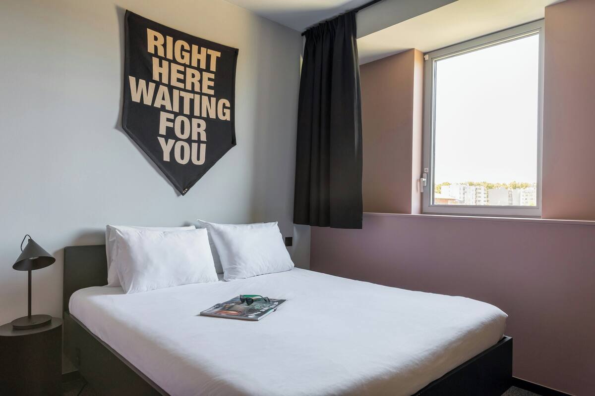 Een hotelkamer met een bed, nachtkastje, lamp, raam met gordijnen en een inspirerend bord aan de muur.