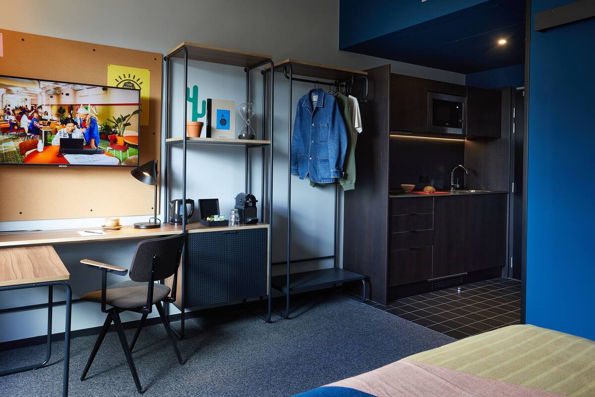 Een compacte hotelkamer met een kitchenette, een bureau met stoel, een bed en een televisie waarop een scène van mensen te zien is die aan het werk zijn.