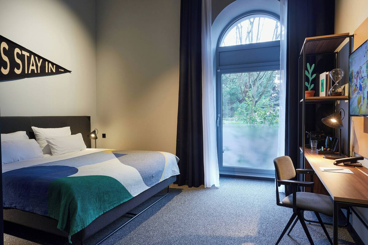 Hotelkamer met een bed, bureau, stoel, boekenkast, groot raam met gordijnen en een bord aan de muur.