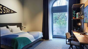 A hotel room with a bed, a desk, a chair, a bookshelf, a large window with curtains, and a sign on the wall.