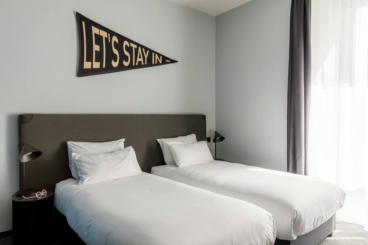 Een hotelkamer met twee bedden, een nachtkastje met een lamp, en een bord aan de muur met de tekst 'LET'S STAY IN'.