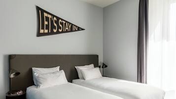 A hotel room with two beds, a nightstand with a lamp, and a wall sign that reads 'LET'S STAY IN.'