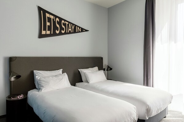 Habitación de hotel con dos camas, una mesita de noche con lámpara y un cartel en la pared que dice 'LET'S STAY IN'.