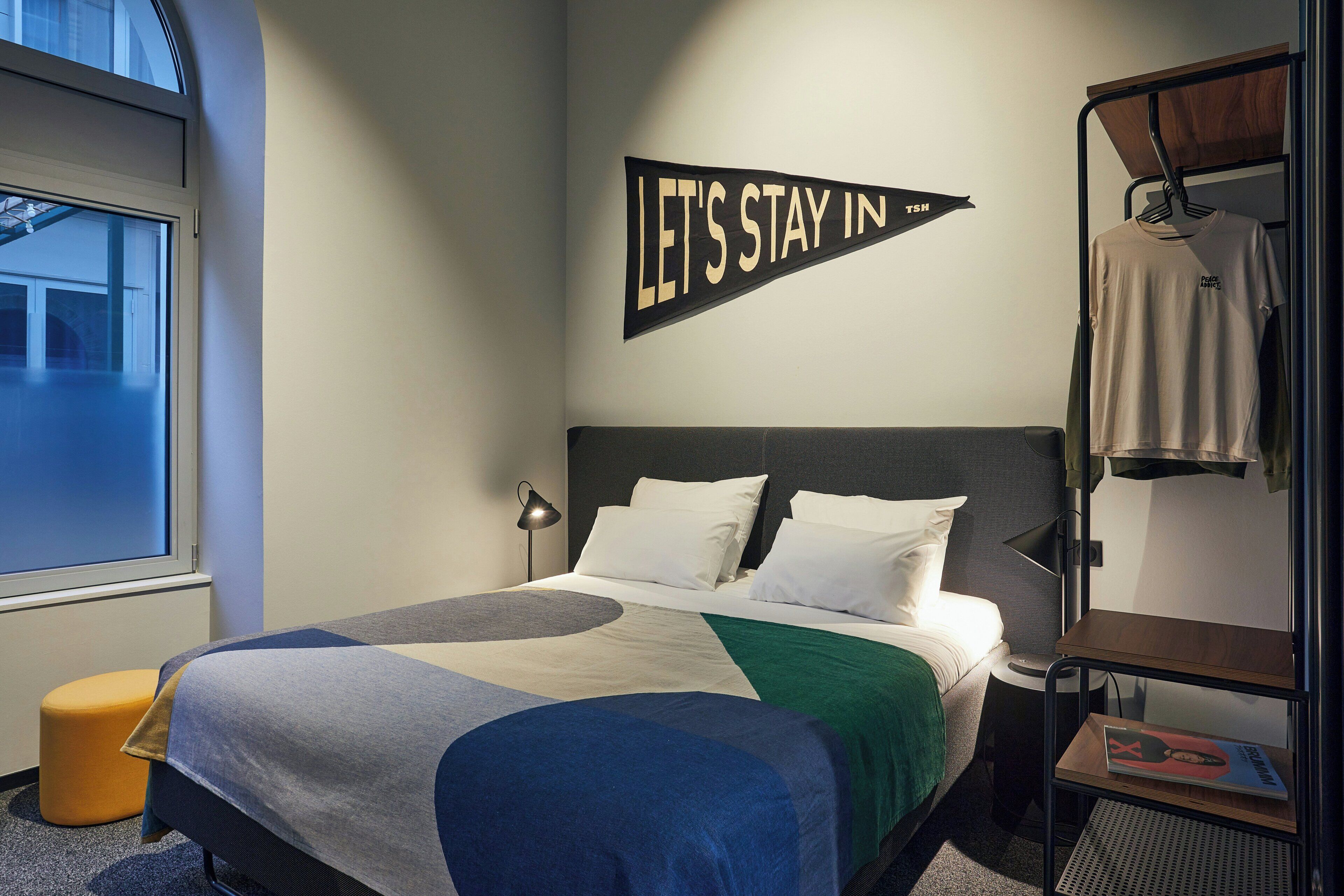 Una cama bien hecha con una colcha de estampado geométrico, un letrero en la pared que dice 'LET'S STAY IN TSH', un perchero con una camiseta y una pequeña mesa de madera con una revista.