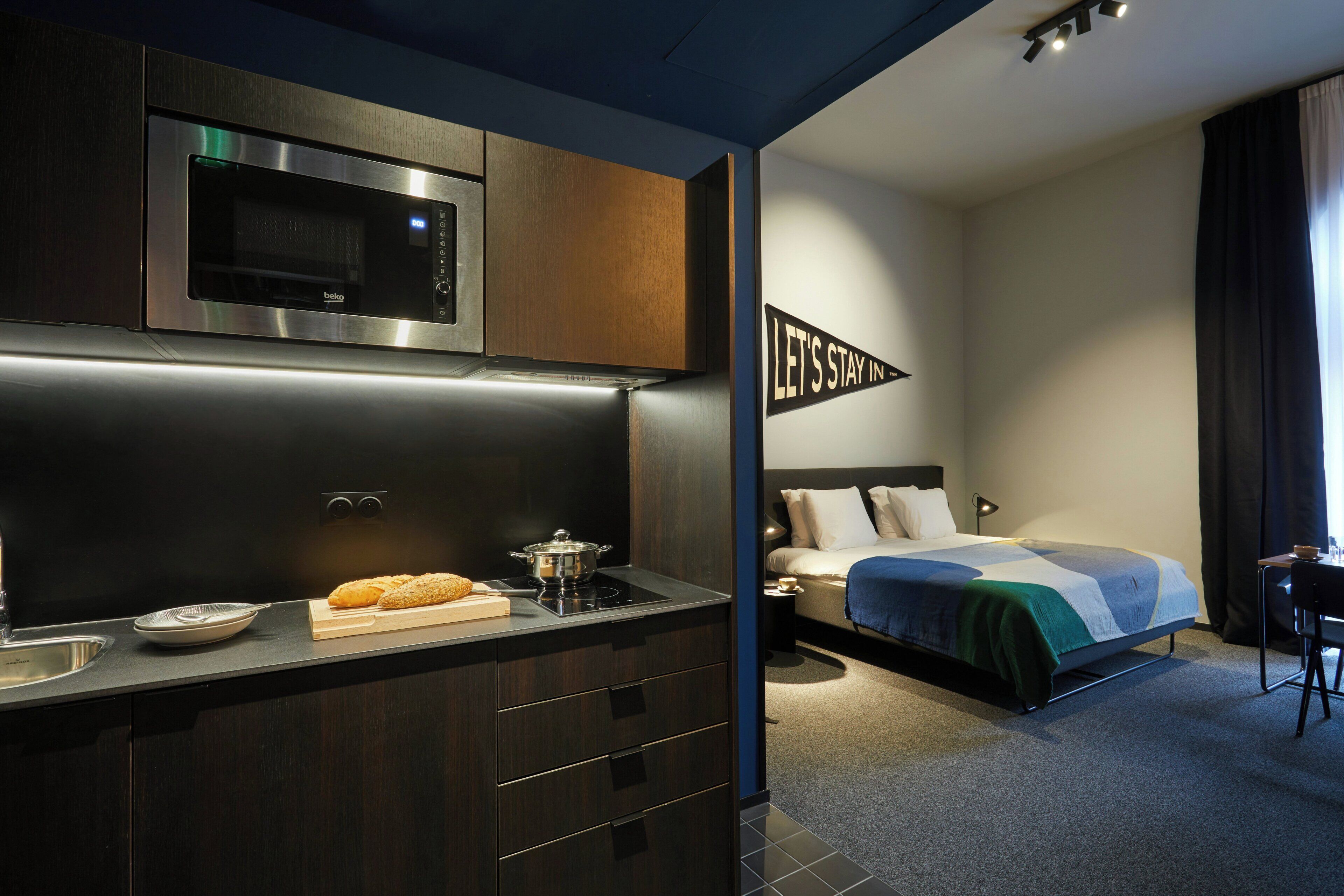 A modern hotel room with a kitchenette, a bed with a blue and white bedspread, a desk, and a nightstand.