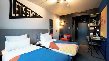 A hotel room with two beds, a desk, a chair, and a bookshelf.