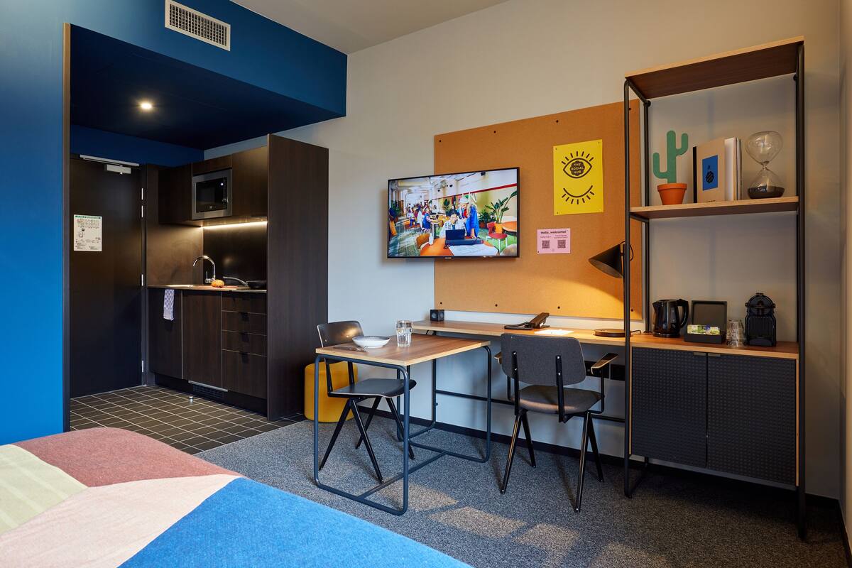 Een moderne hotelkamer met een bureau, twee stoelen, een televisie, een klein plankje met decoratieve spullen, en een bed met een kleurrijke deken.