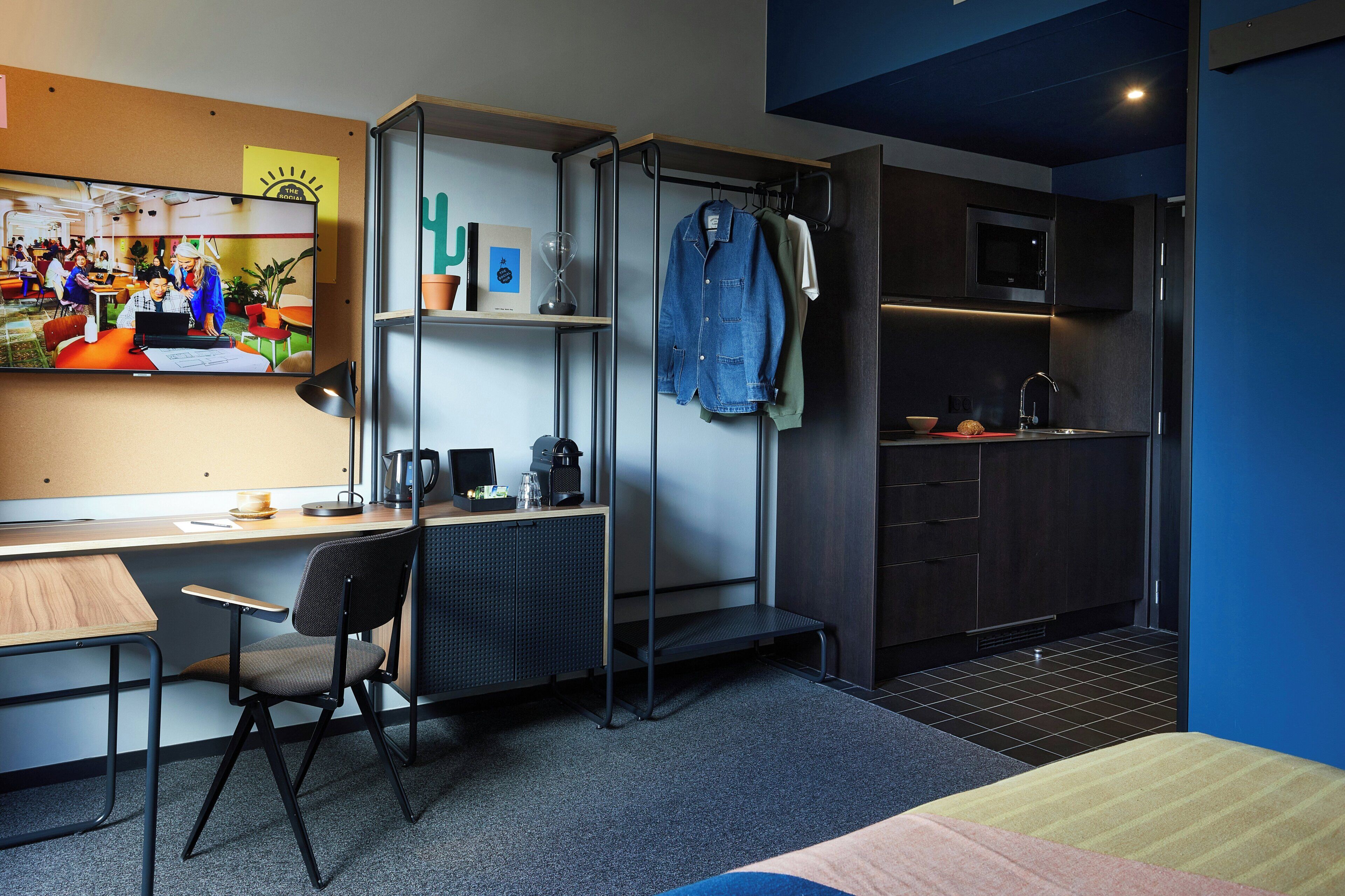 A modern room with a desk, chair, kitchenette, and a TV displaying a group of people.