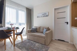 A living room with a sofa, a dining table with chairs, a painting on the wall, and a door.
