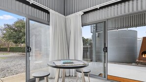 A modern outdoor patio with a round table and chairs, a glass door leading to a garden, and a rustic fire feature. - The Cubby House by Wine Coast Holiday Rentals (WHITES VALLEY)