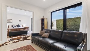 A modern living room with a leather sofa, a wooden bed, and a large window offering a view of a hedge and sky. - Hideaway Nook by Wine Coast Holiday Rentals (WHITES VALLEY)