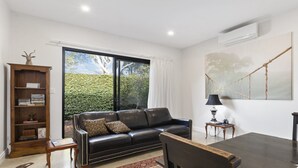 A modern living room with a leather sofa, a wooden bookshelf, and a large window with a view of greenery. - Hideaway Nook by Wine Coast Holiday Rentals (WHITES VALLEY)