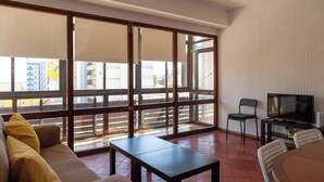 A spacious living area with a sofa, a coffee table, and a TV on a stand. Large windows offer a view of the cityscape.