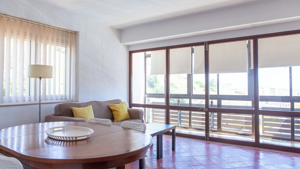 A spacious room with a large window, a wooden table, a sofa with cushions, and a lamp.