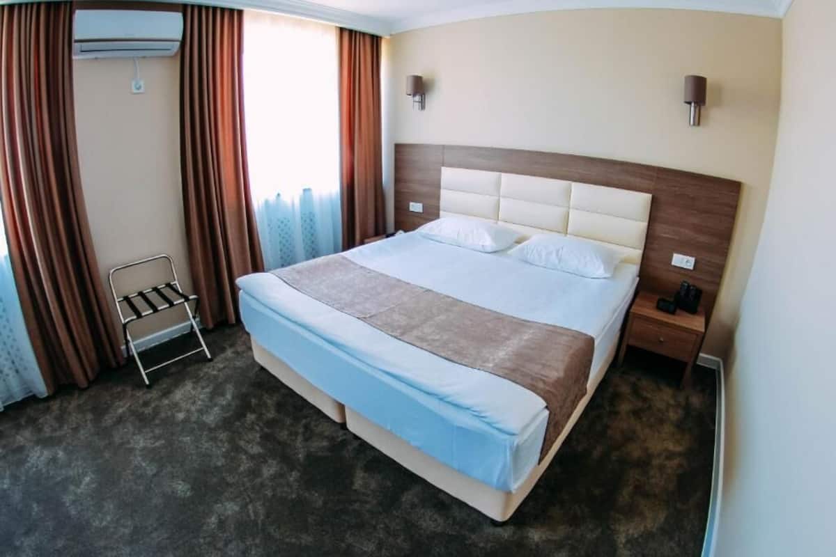 A hotel room with a large bed, a bedside table, an air conditioning unit, and a window with curtains.