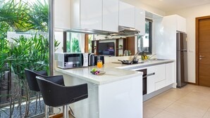 A modern kitchen with white cabinets, a stainless steel refrigerator, a microwave, and a sink. There are two black bar stools and a glass partition with plants. - Two Storey Villa Kael in KA Villas Rawai (Rawai)