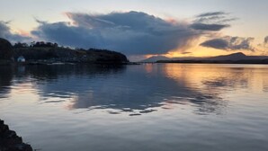 Um lago tranquilo ao pôr do sol, com uma cadeia montanhosa distante e uma pequena casa na margem.