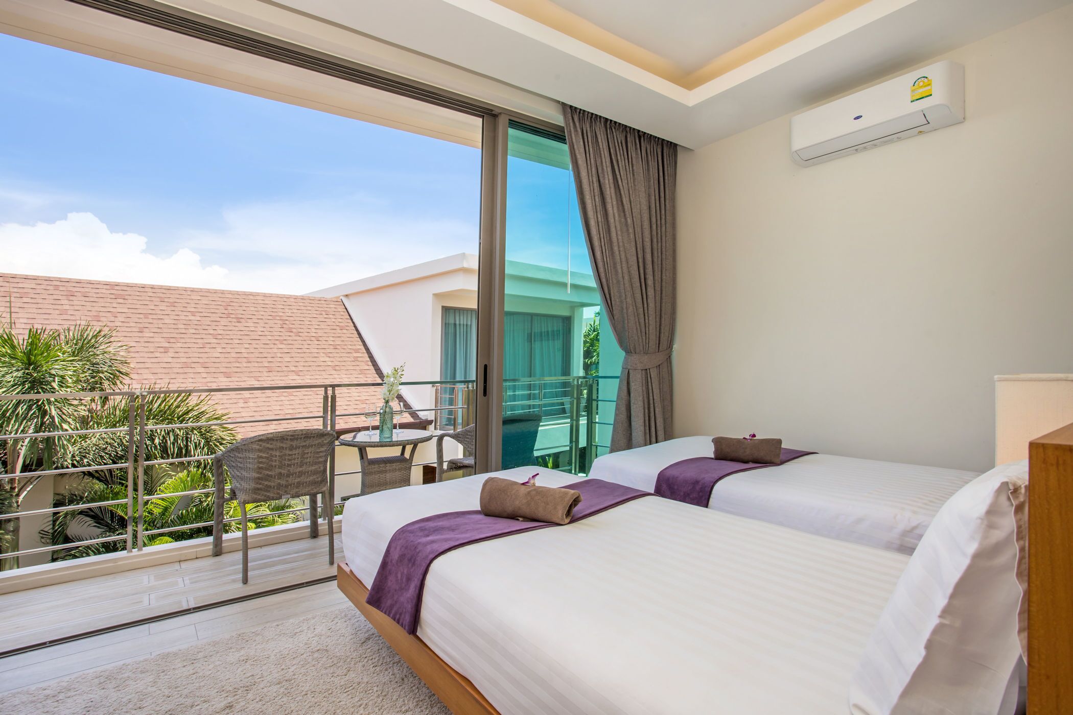 A hotel room with two beds, a balcony with a table and chairs, and a view of a building and greenery.
