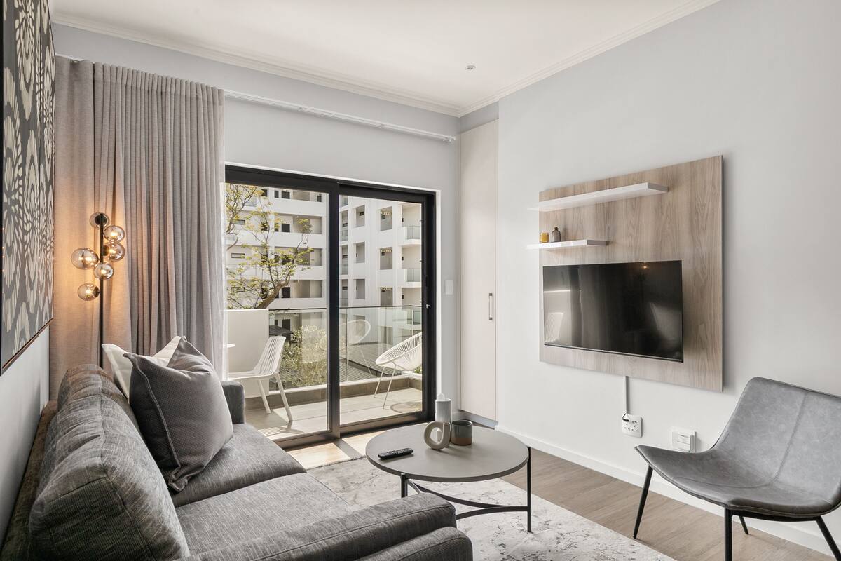 A modern living room with a grey sofa, a round coffee table, and a grey armchair. There is a flat-screen TV mounted on the wall, a large window with a view of a building, and a balcony with a chair.
