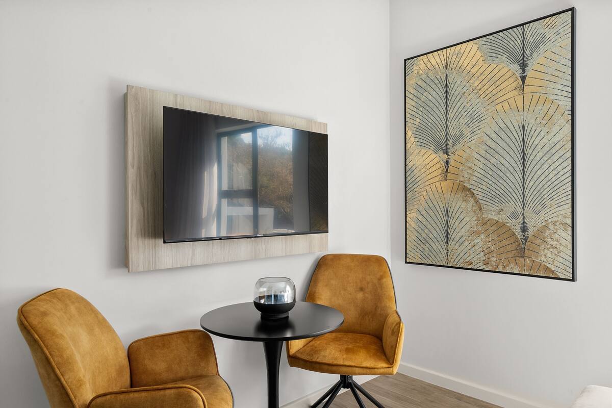 A modern room with a large flat-screen TV mounted on a wooden panel, two mustard-colored armchairs around a small round table, and a large abstract painting on the wall.