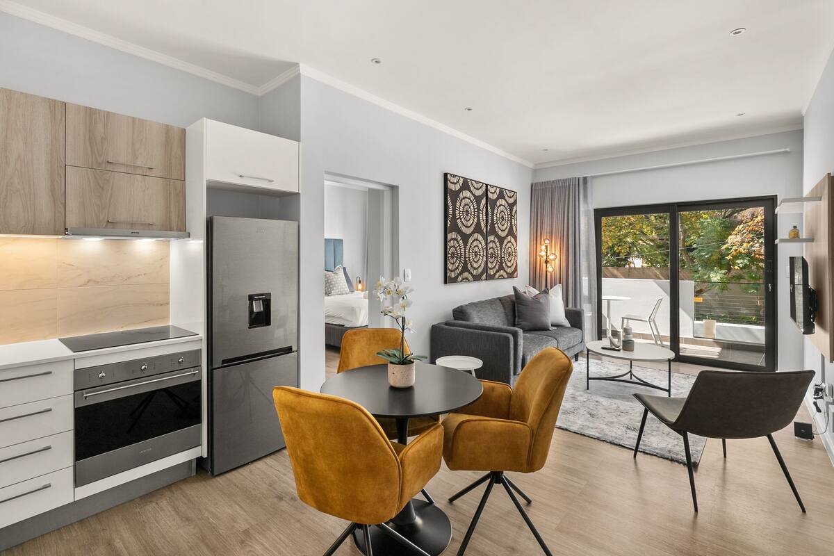 A modern kitchen with built-in appliances, a dining area with a round table and chairs, and a living space with a sofa and a large window.