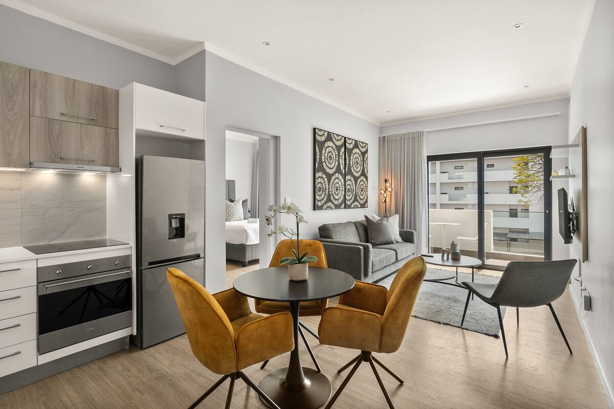 A modern kitchen with built-in appliances, a dining area with a round table and chairs, and a living space with a sofa and a large window.